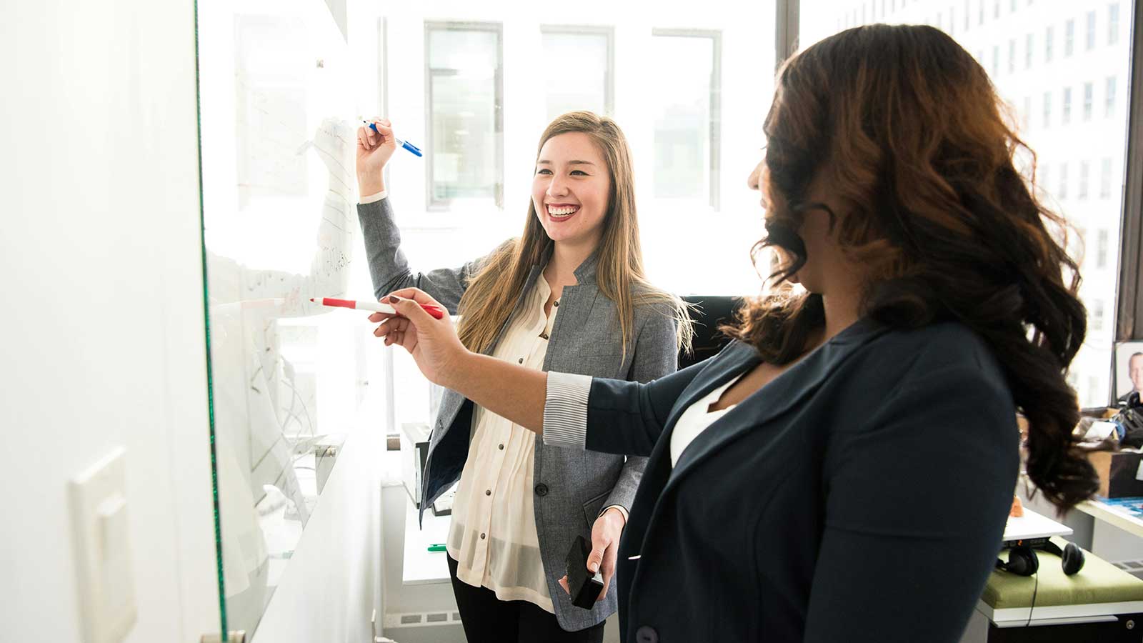 Two colleagues working through a diagram on a glass office wall beside a bright window