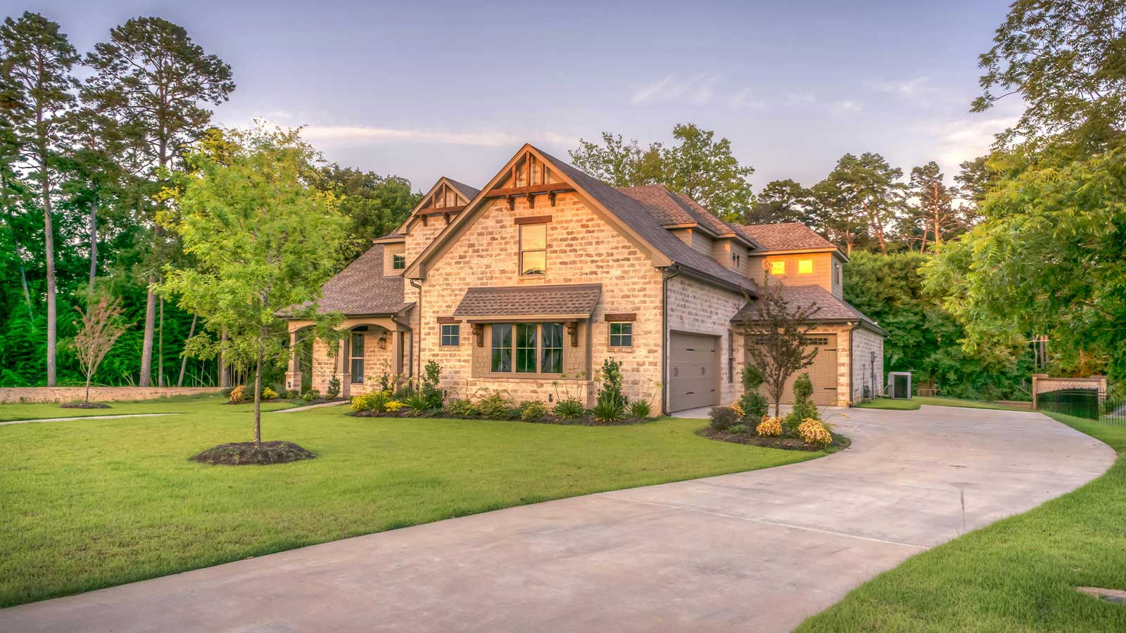 A large stone faced house at dusk with a curving concrete drive and mature trees around it