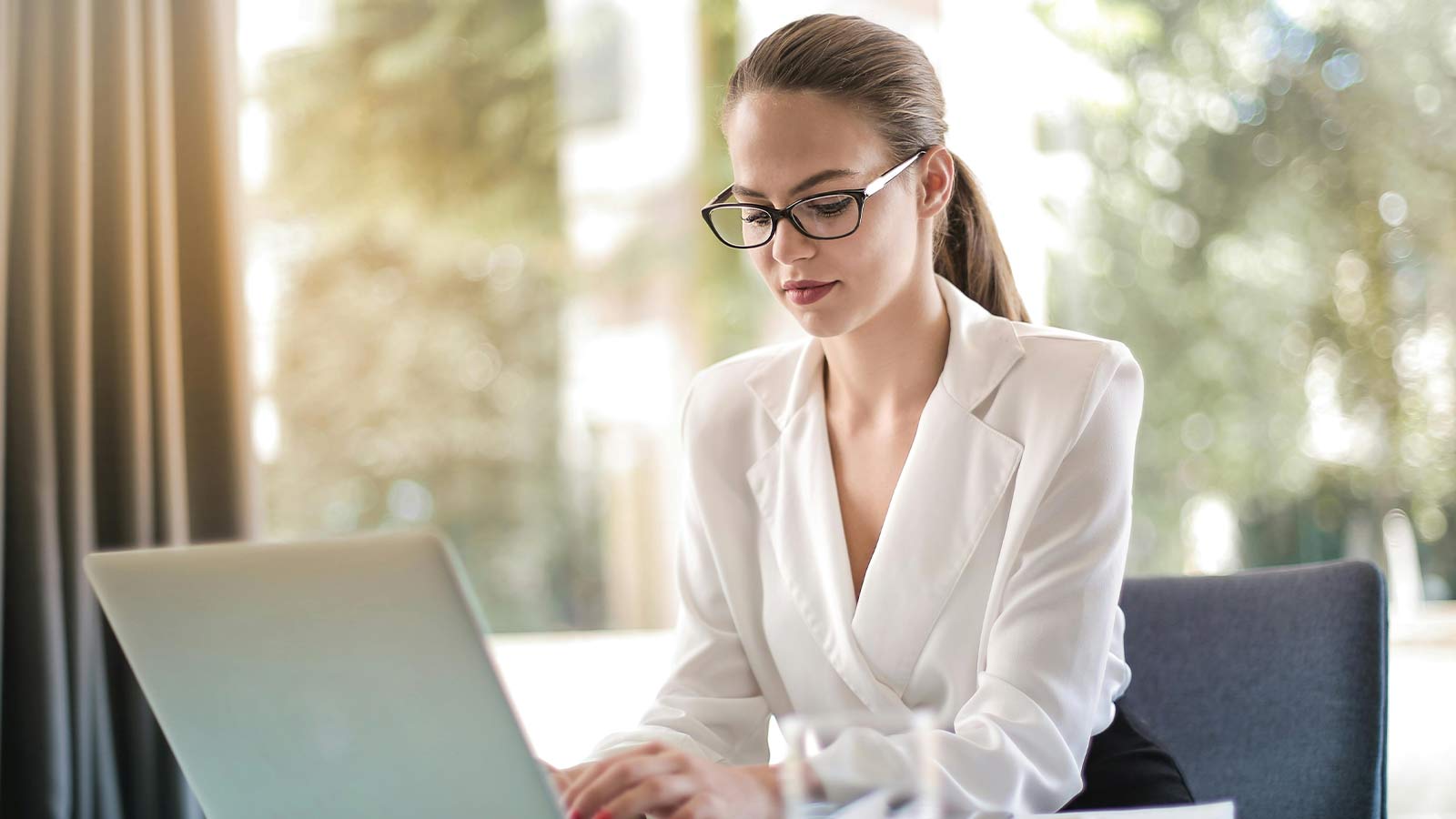 A professional working at a laptop beside a bright window with trees outside