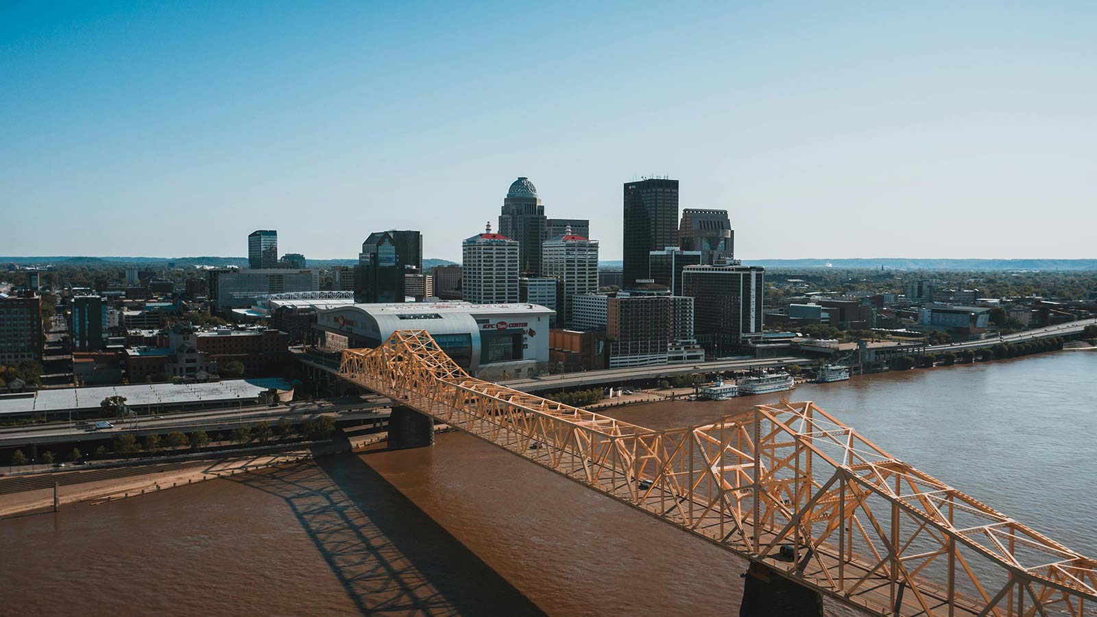 A yellow steel truss bridge crossing a wide brown river toward a riverfront city skyline