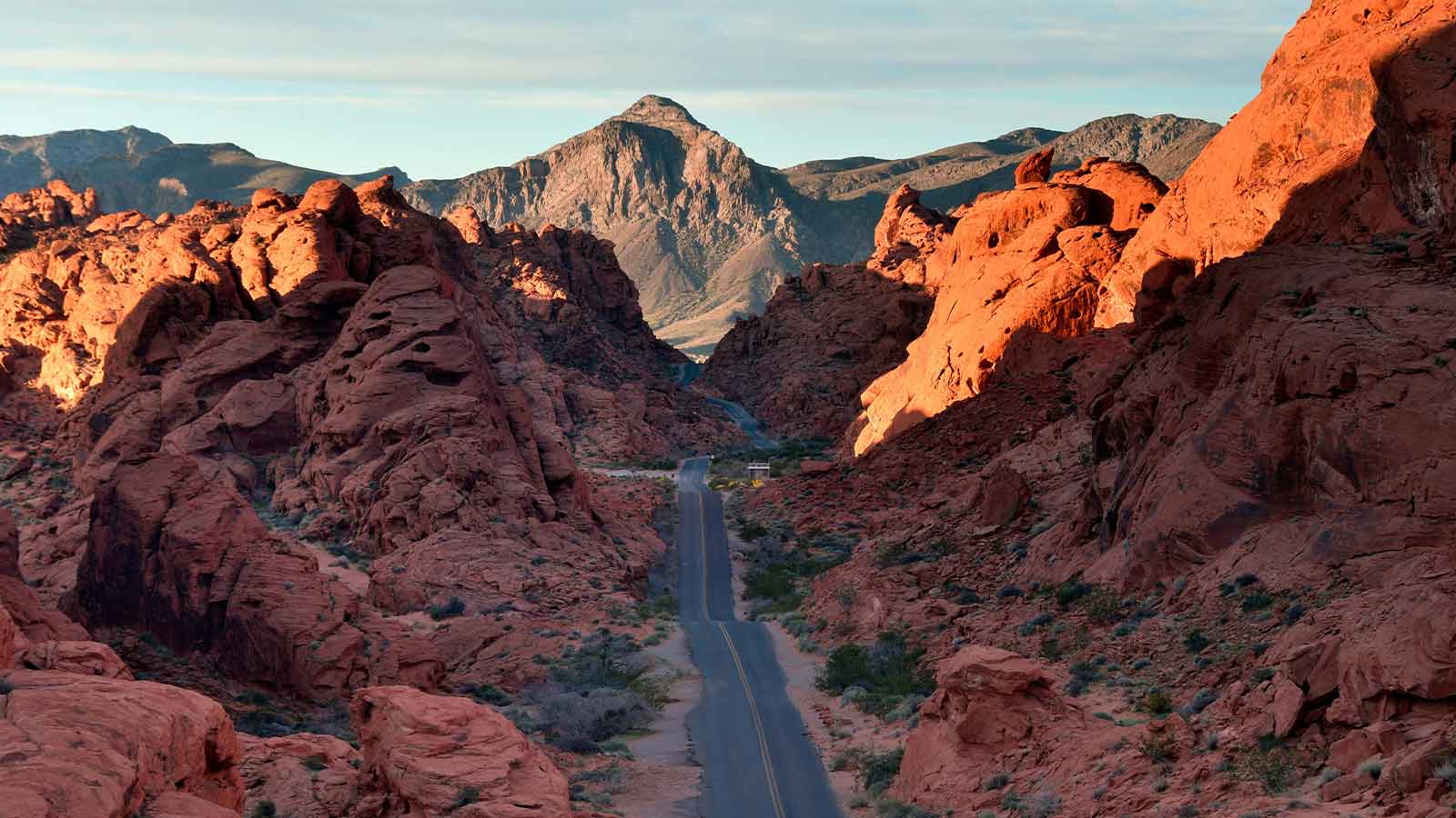 A two lane road running between red sandstone formations toward a grey desert mountain at sunrise in southern Nevada