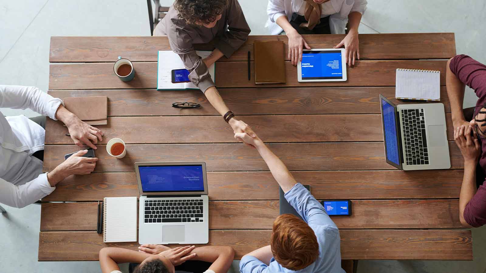 Five people meeting around a wooden table with laptops and notebooks seen from above