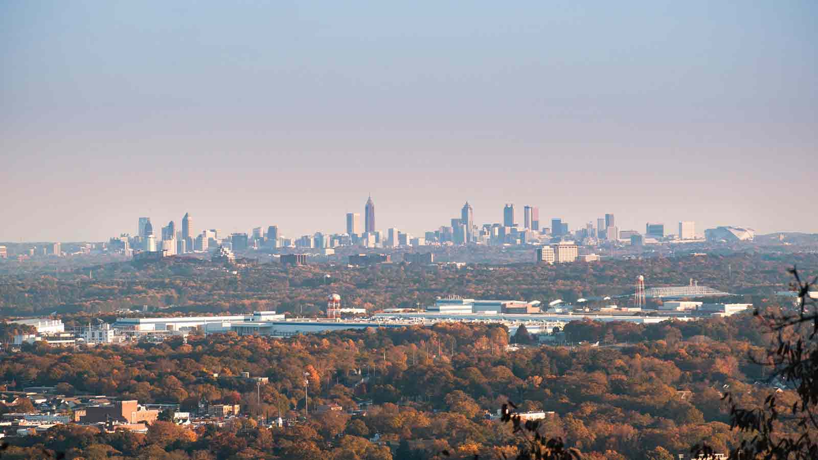 A hazy city skyline seen from a distance across miles of autumn tree canopy at golden hour